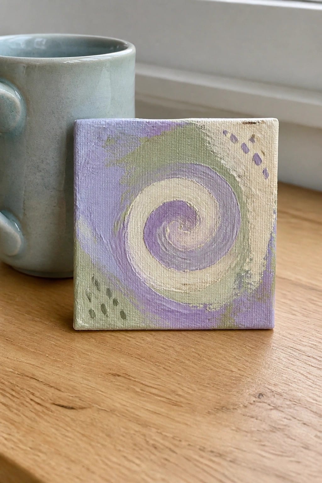 Small square canvas with purple spiral beside light blue ceramic mug on wood
