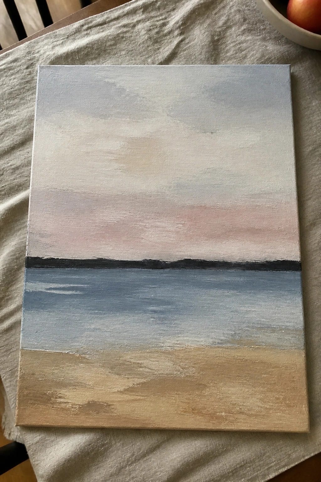 A rectangular oil painting showing a soft gradient sky over a dark horizon line, blue middle section, and beige bottom area on canvas resting on fabric.