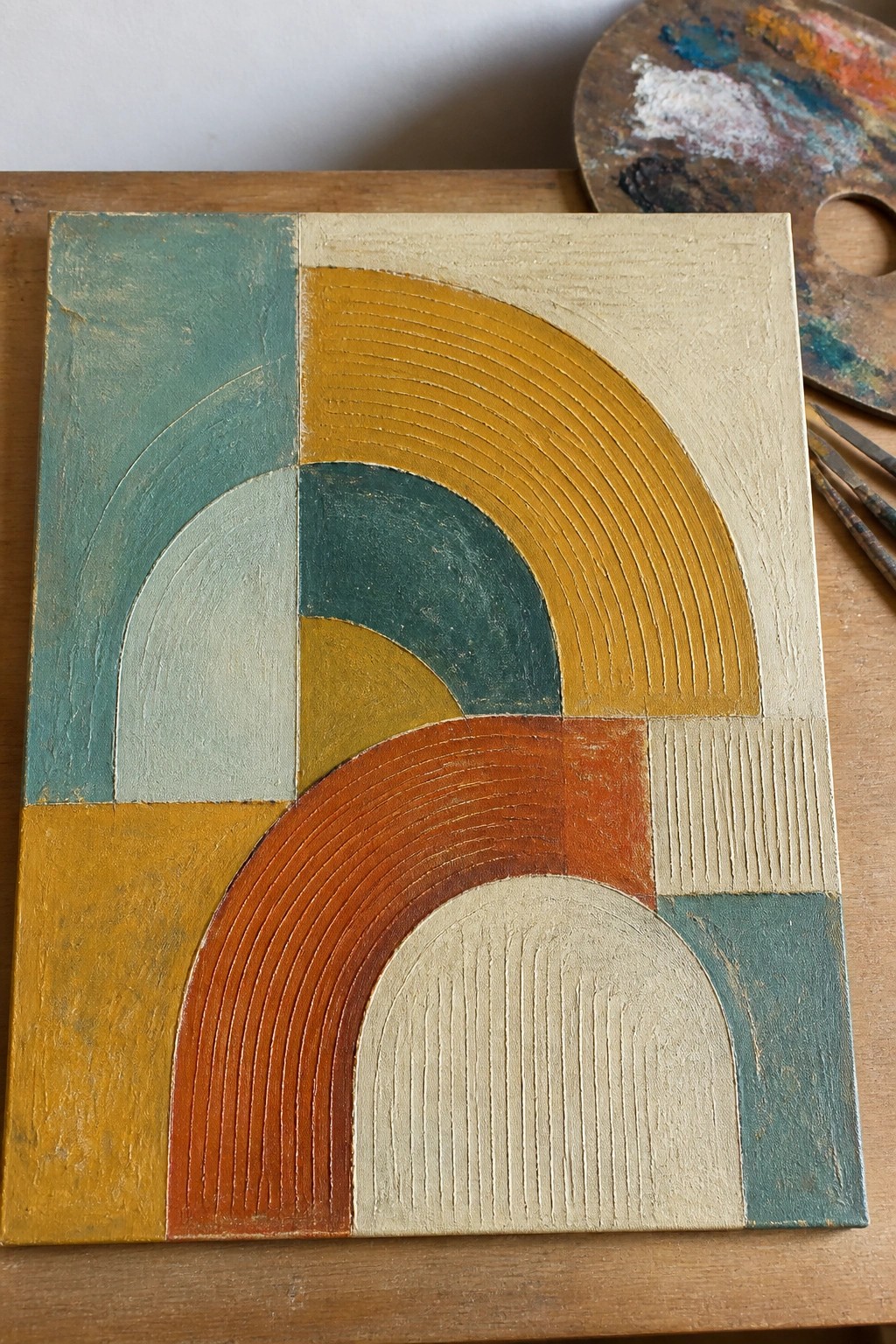 Abstract artwork with colorful geometric arches beside paint palette and brushes on wood.
