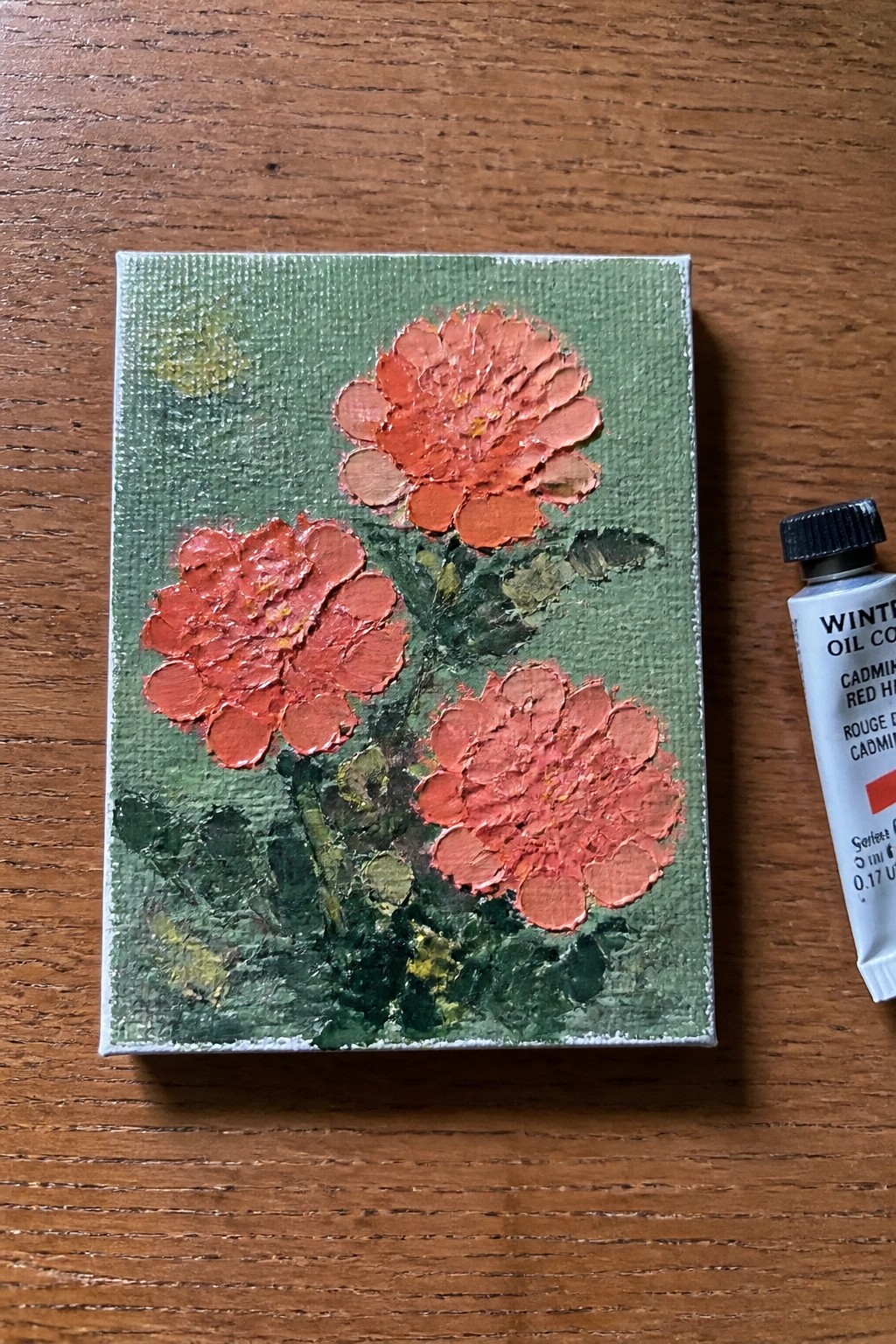 Small canvas with impasto red flowers on green beside cadmium red oil paint tube on wood