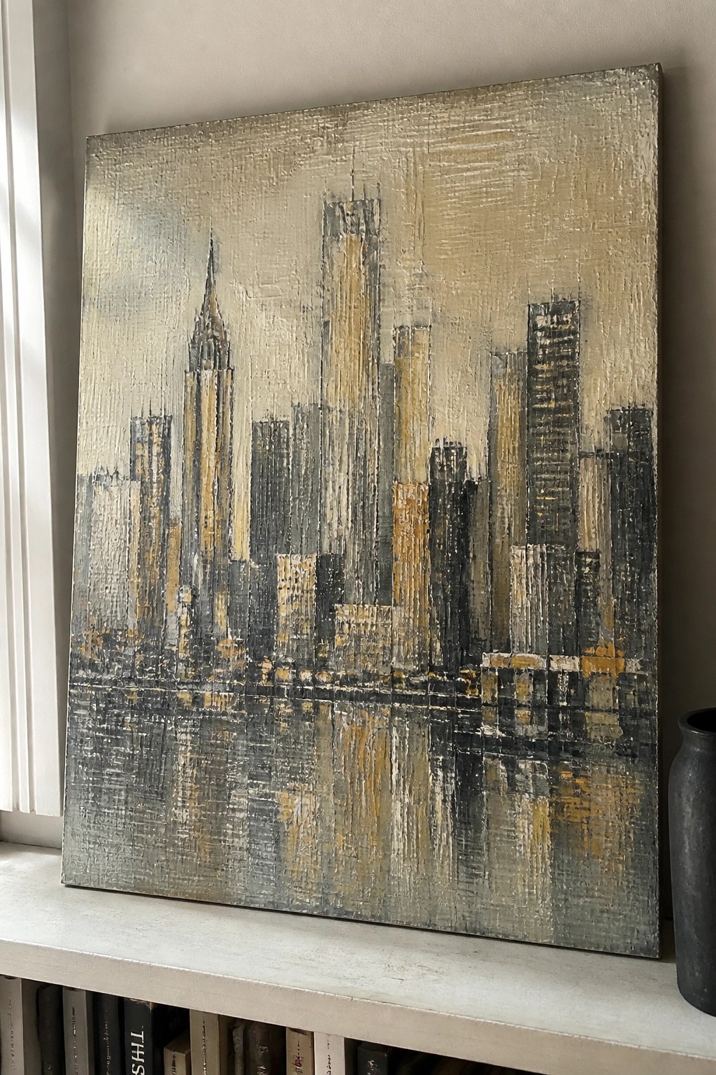 Abstract city skyline painting with water reflections in muted grays and golds on shelf.