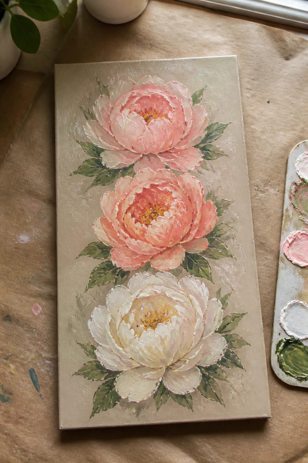 Three pink and white peonies with green leaves on beige canvas beside palette