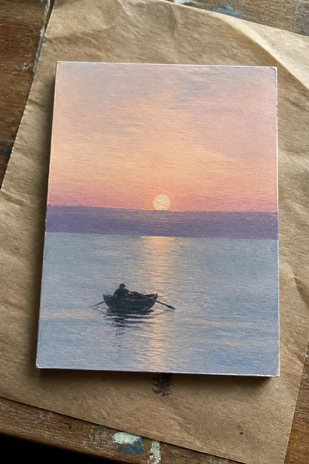 Silhouetted rower in small boat on calm water beneath pink-orange sunset sky with purple horizon