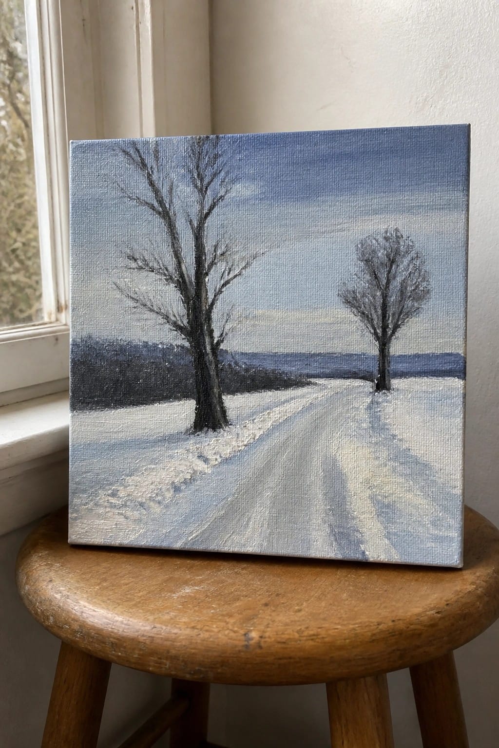 Winter landscape painting of snowy path with two bare trees on wooden stool