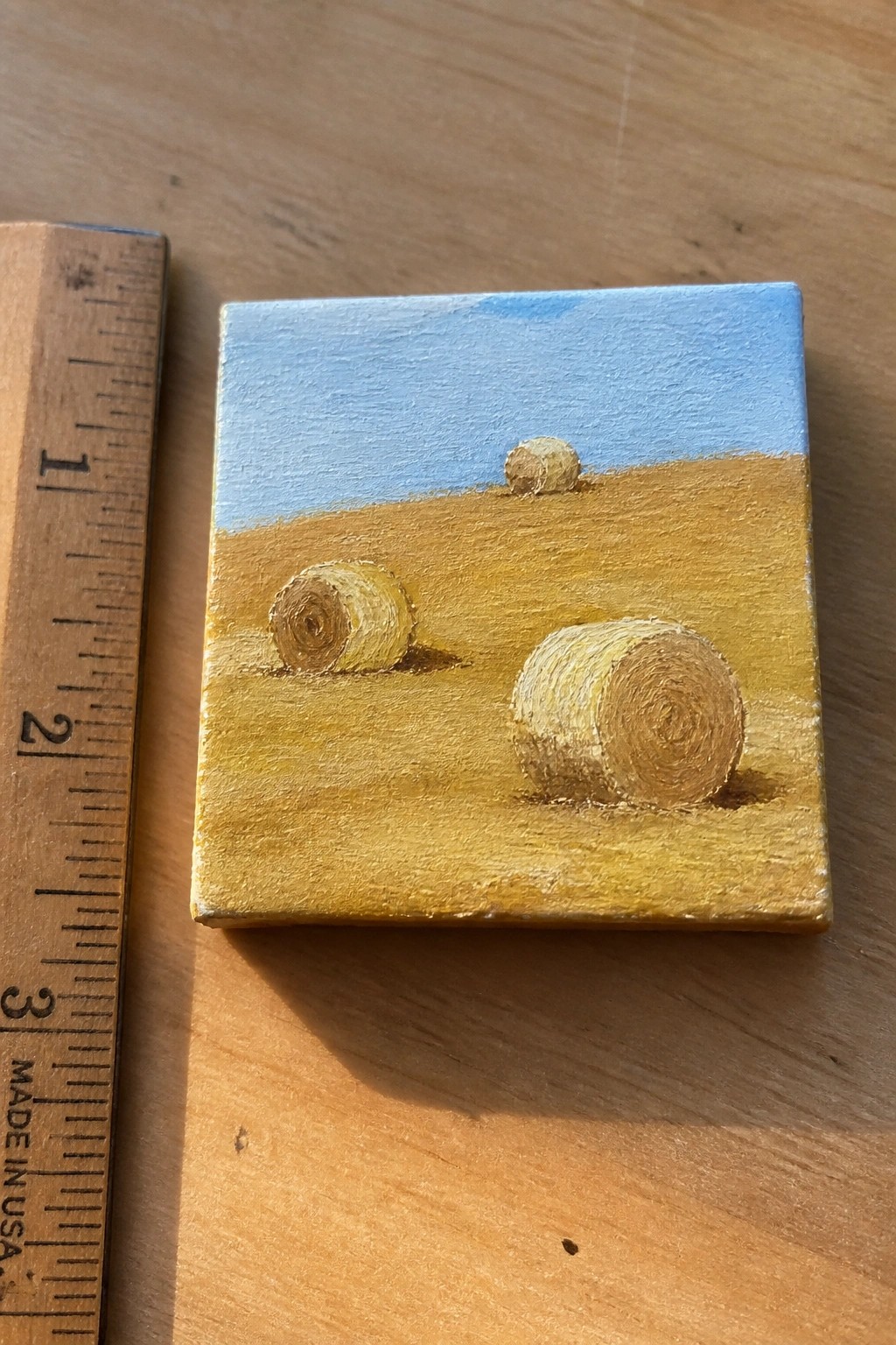 Three hay bales in a golden field painted on small canvas beside wooden ruler