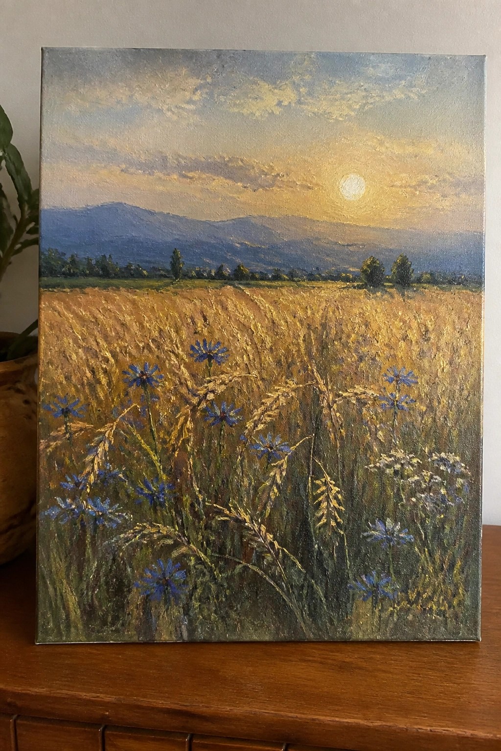 Golden wheat field with blue wildflowers, distant mountains under a glowing sunset in oil painting
