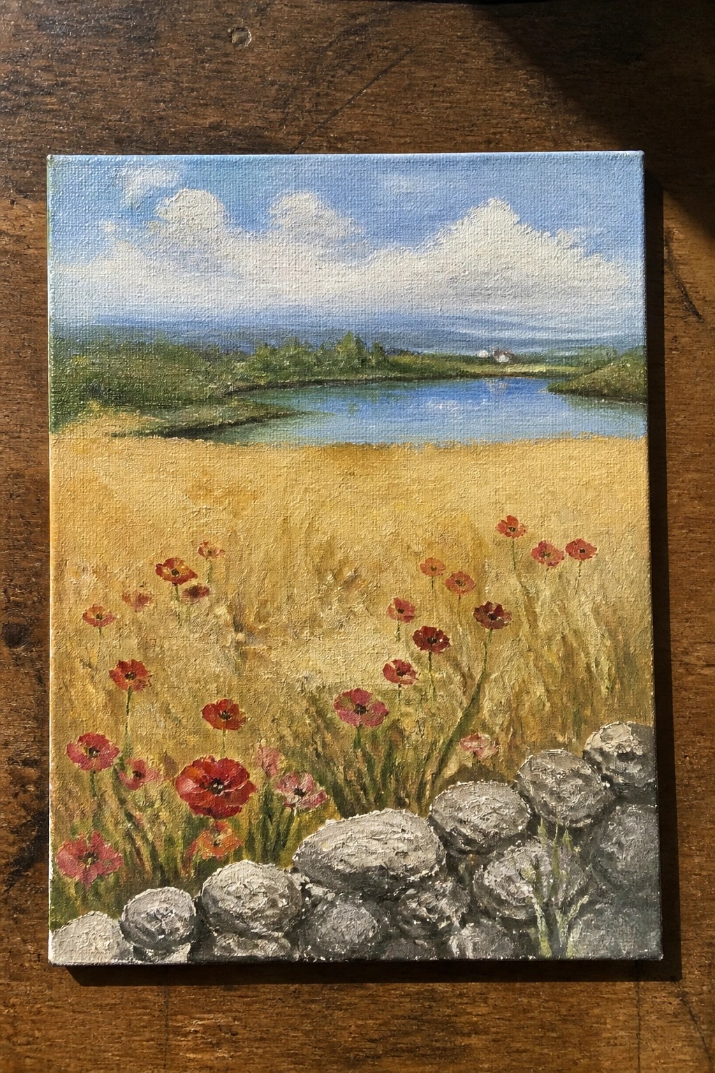 Golden field with red poppies, stone wall, blue river, and cloudy sky.