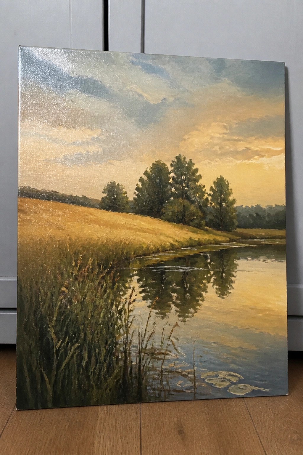An oil painting of a golden field with a reflective river under a warm sunset sky and trees in the background.