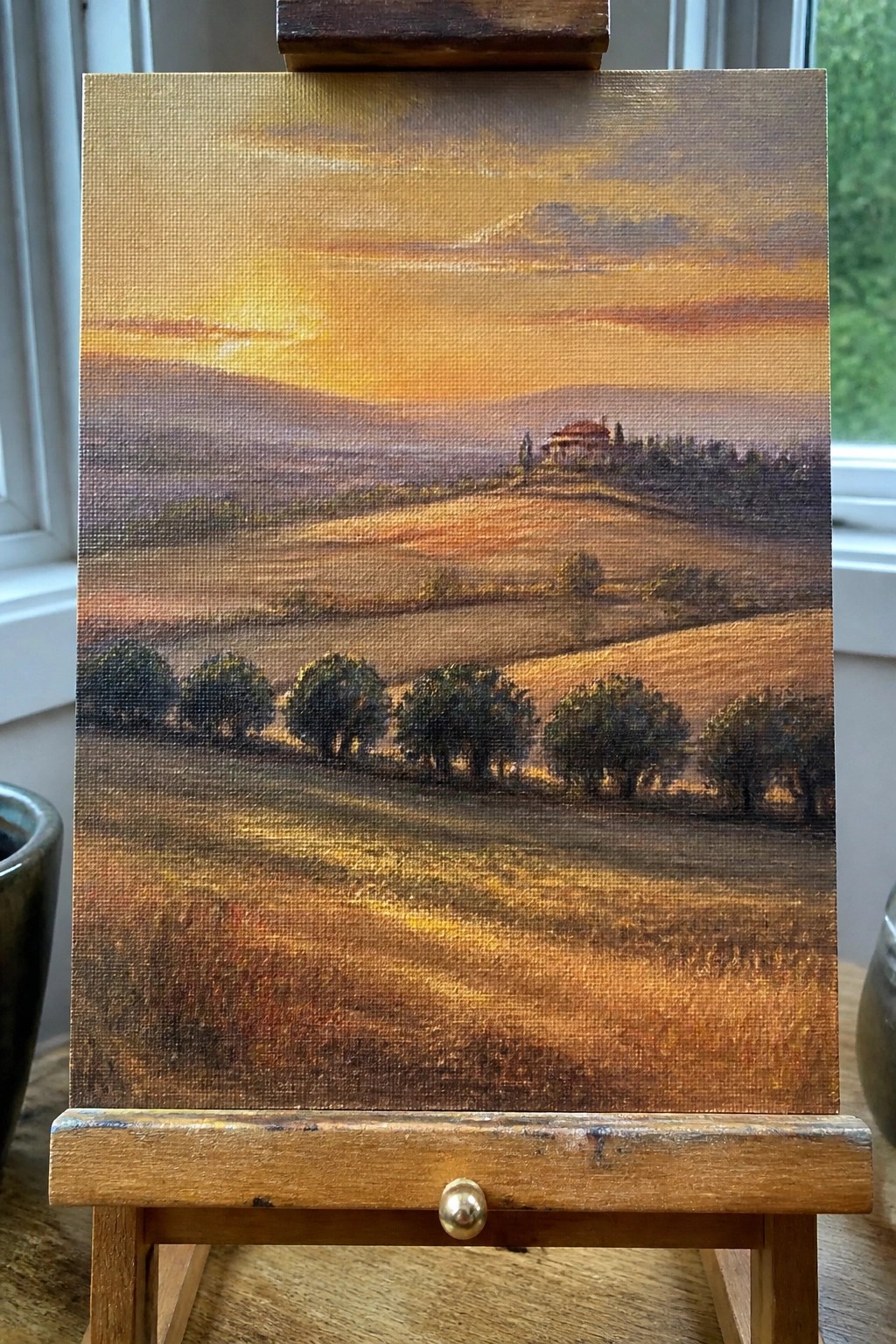 Sunset landscape painting with golden fields, trees, and house on wooden easel.