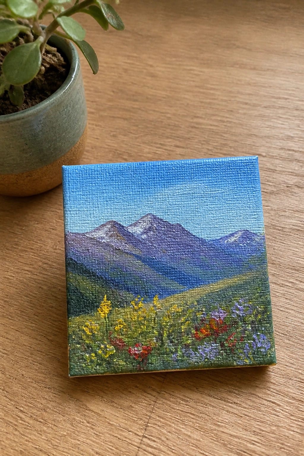 Small canvas painting of purple mountains with colorful wildflowers beside potted plant on wood.