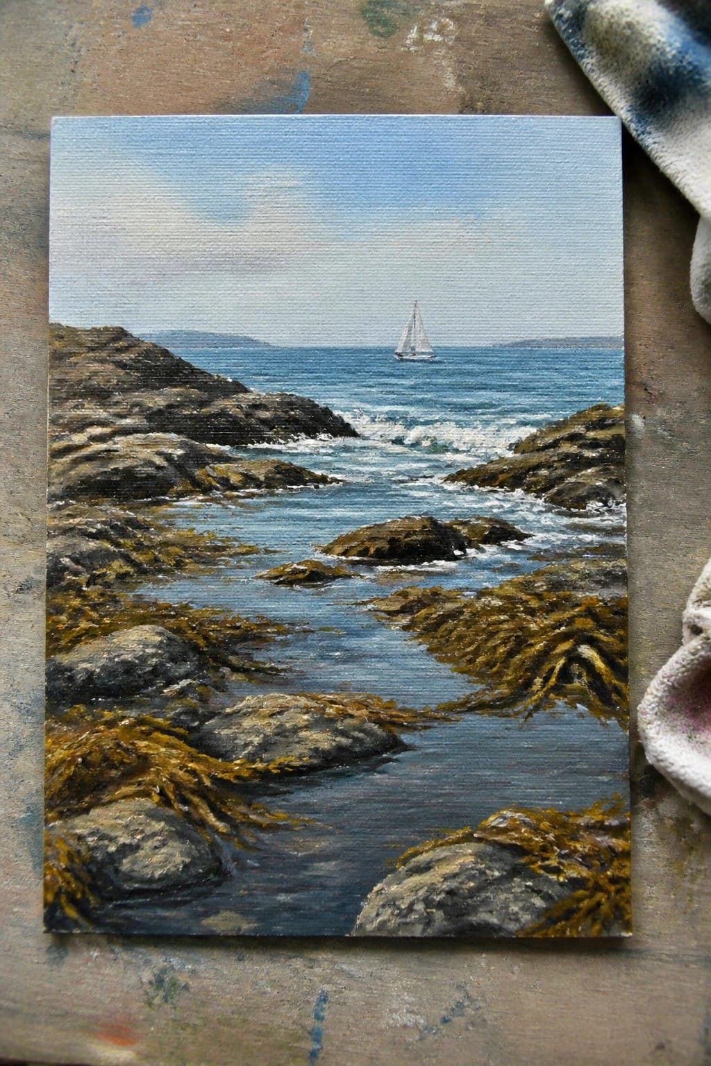Coastal painting of blue ocean waves on rocky shore with brown seaweed and distant sailboat