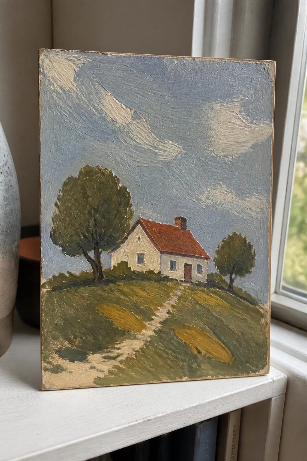 Small painting of white house with red roof on grassy hill with trees and path.