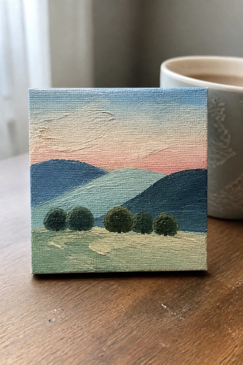 A small square oil painting of rolling blue and green hills with a row of dark green trees in the foreground under a blue-to-pink gradient sky, sitting on a wooden table beside a mug.