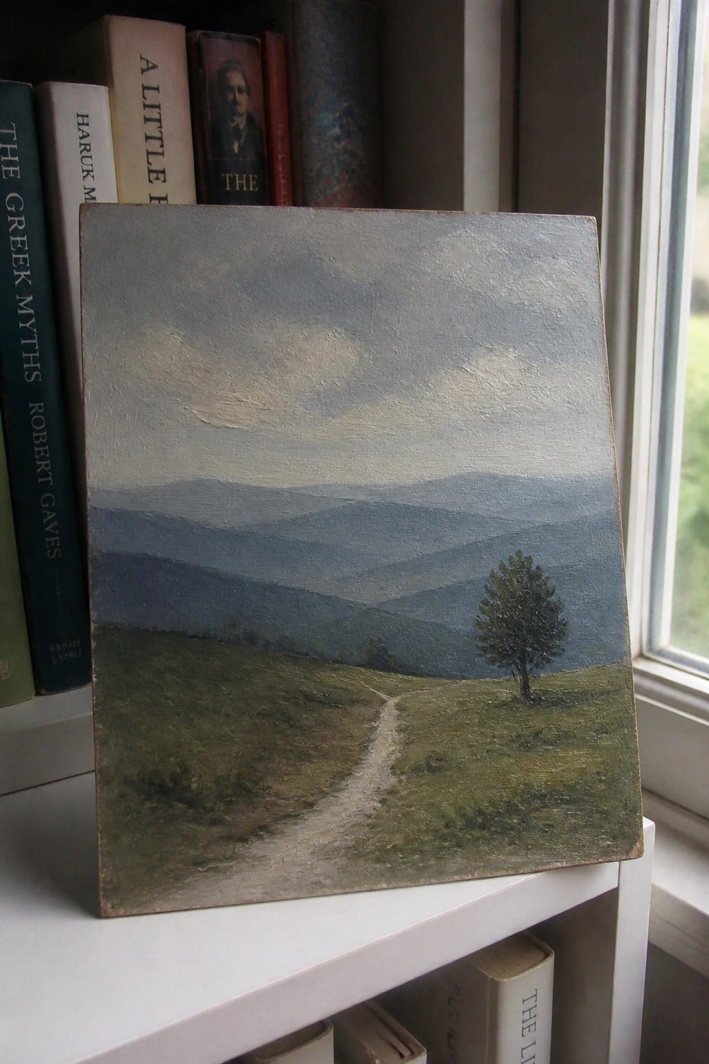 Landscape painting of misty blue hills, winding path, and lone tree on shelf with books