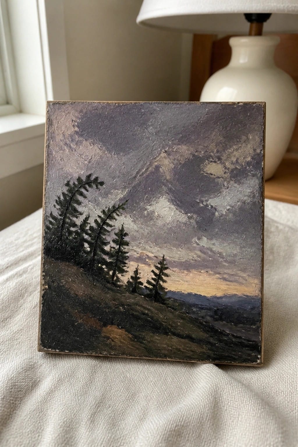 Small impasto oil painting of pine trees and misty mountains on white fabric beside lamp