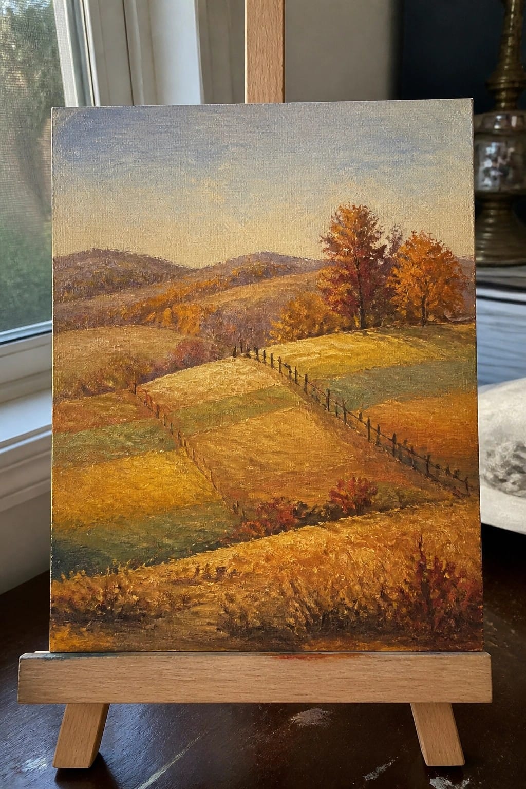 Autumn landscape oil painting with rolling fields, orange trees, and fence on wooden easel