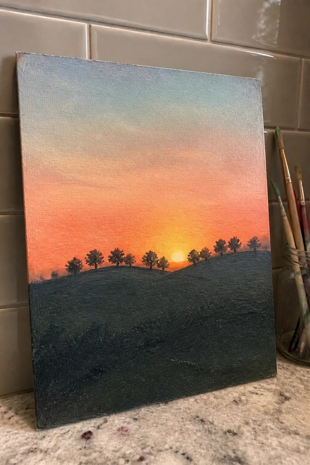 Canvas painting of vibrant sunset over silhouetted trees on dark rolling hills beside brushes