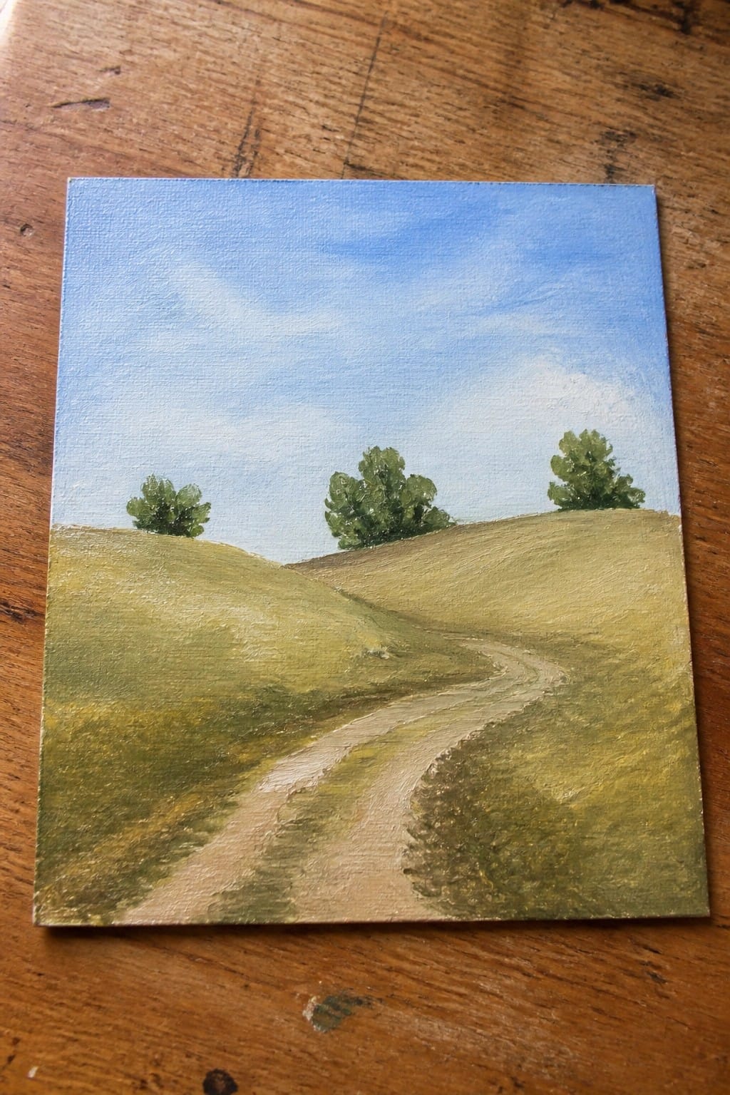 Winding dirt path through rolling green hills with trees under blue sky on wood