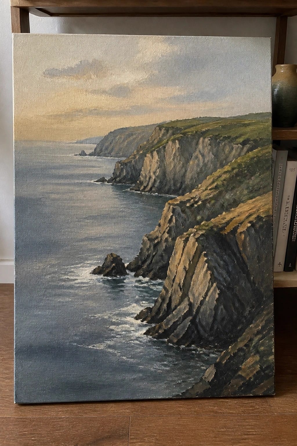 Oil painting of rugged cliffs meeting the sea under a golden sunset sky beside books.