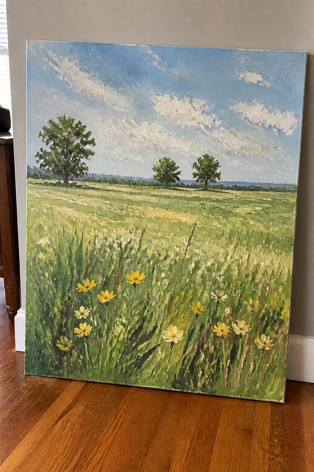 Oil painting of green meadow with yellow wildflowers, trees, and blue sky with clouds.