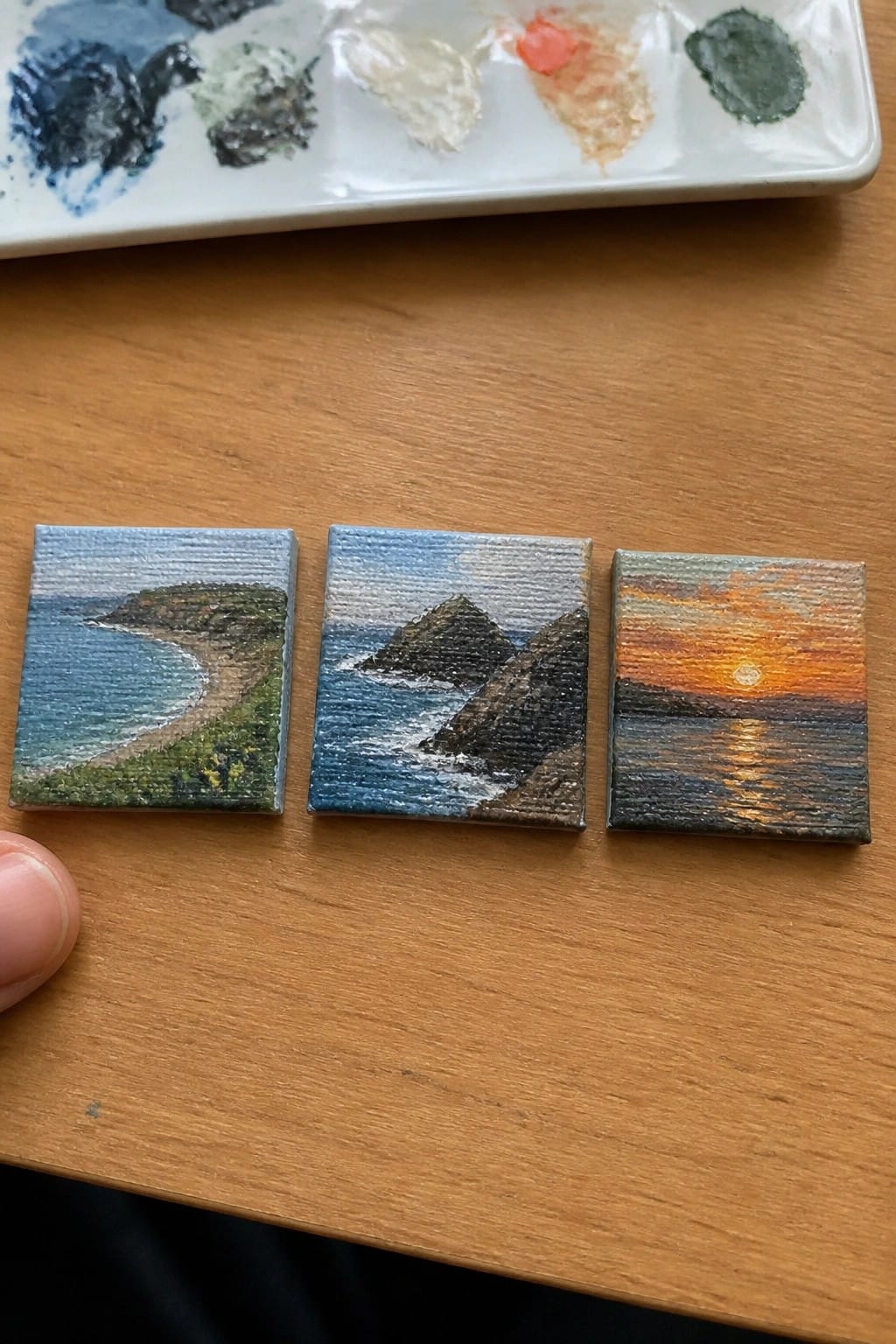 Three miniature seascape paintings on wood beside a paint palette and finger