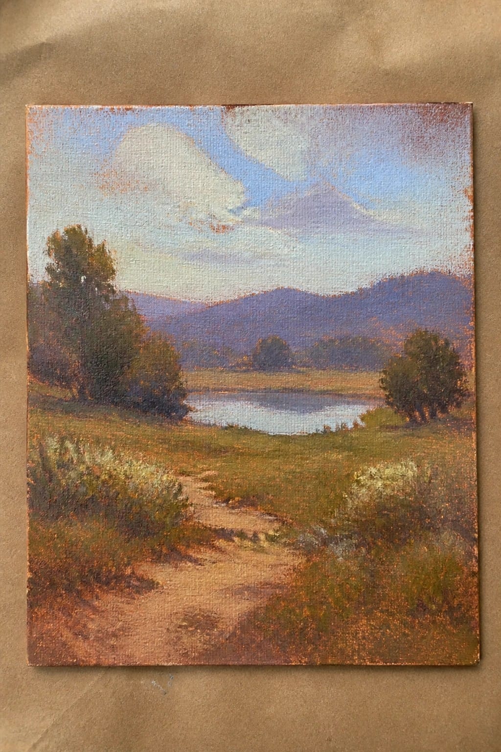 Oil painting of winding dirt path to lake, trees, and distant mountains under clouds.
