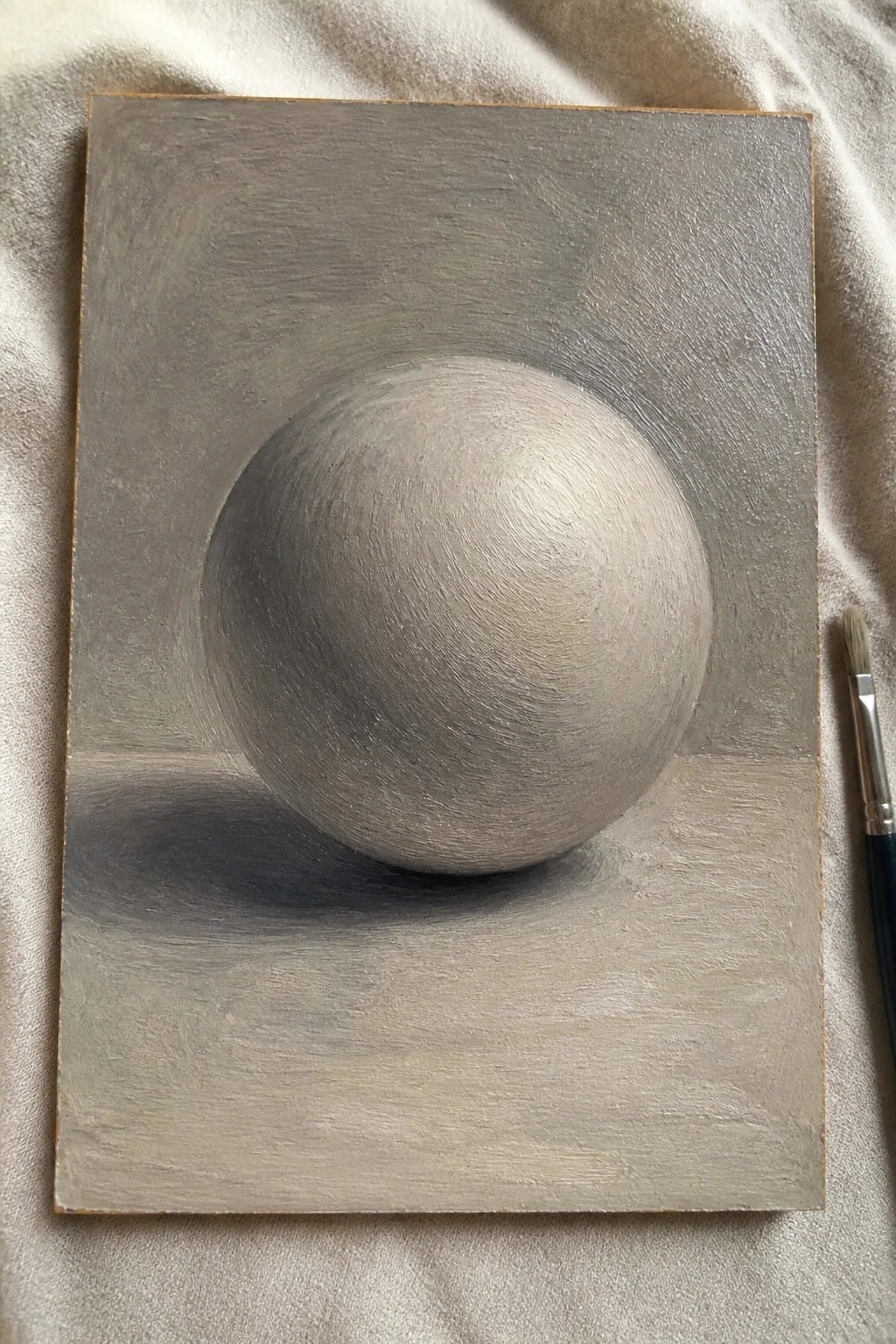 Gray sphere painting with soft shadows on canvas beside paintbrush on white fabric
