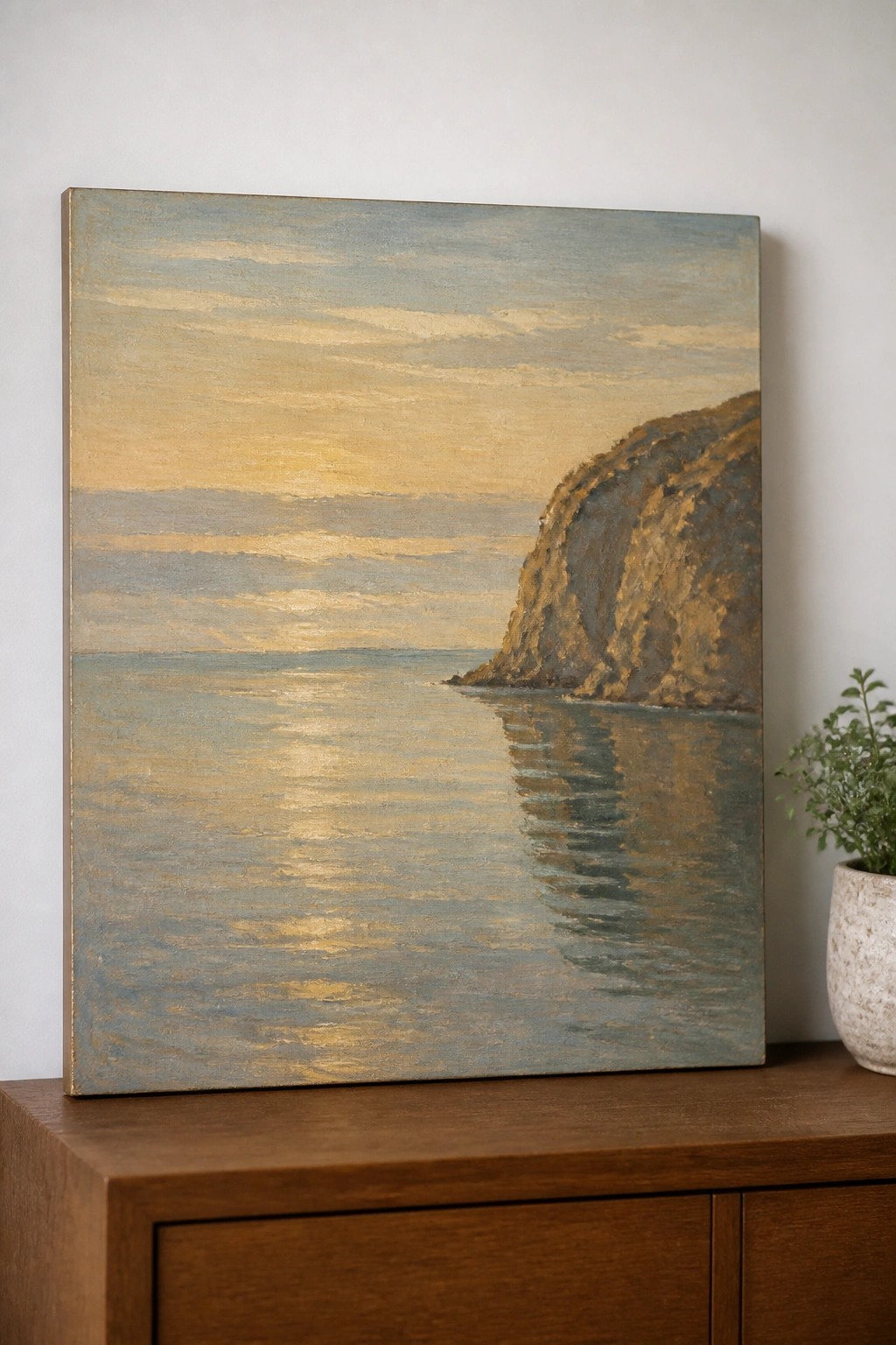 Sunset seascape painting with golden water reflections and rocky cliff on wooden cabinet beside plant.