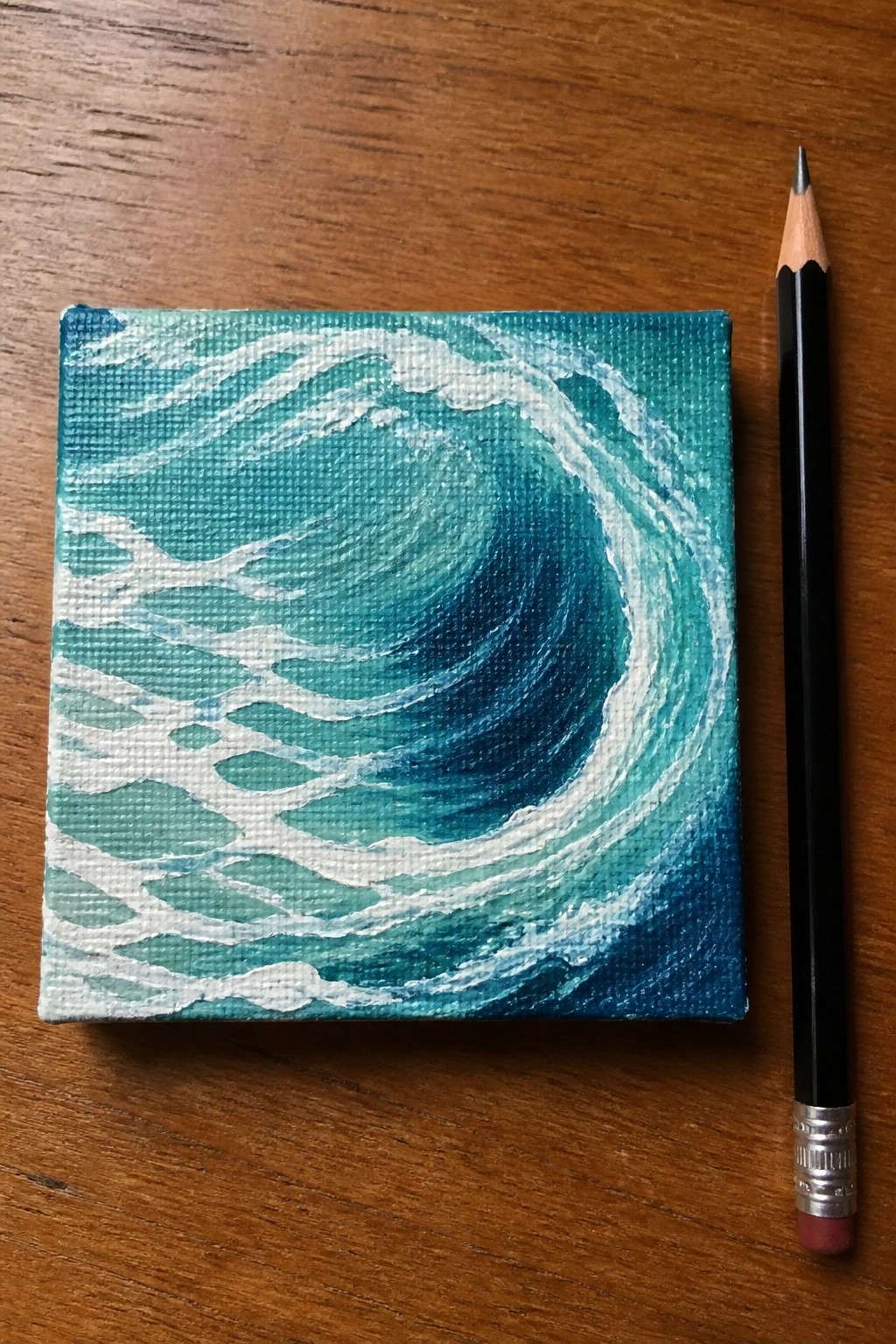 Small square canvas with blue curling wave painting beside black pencil on wood.