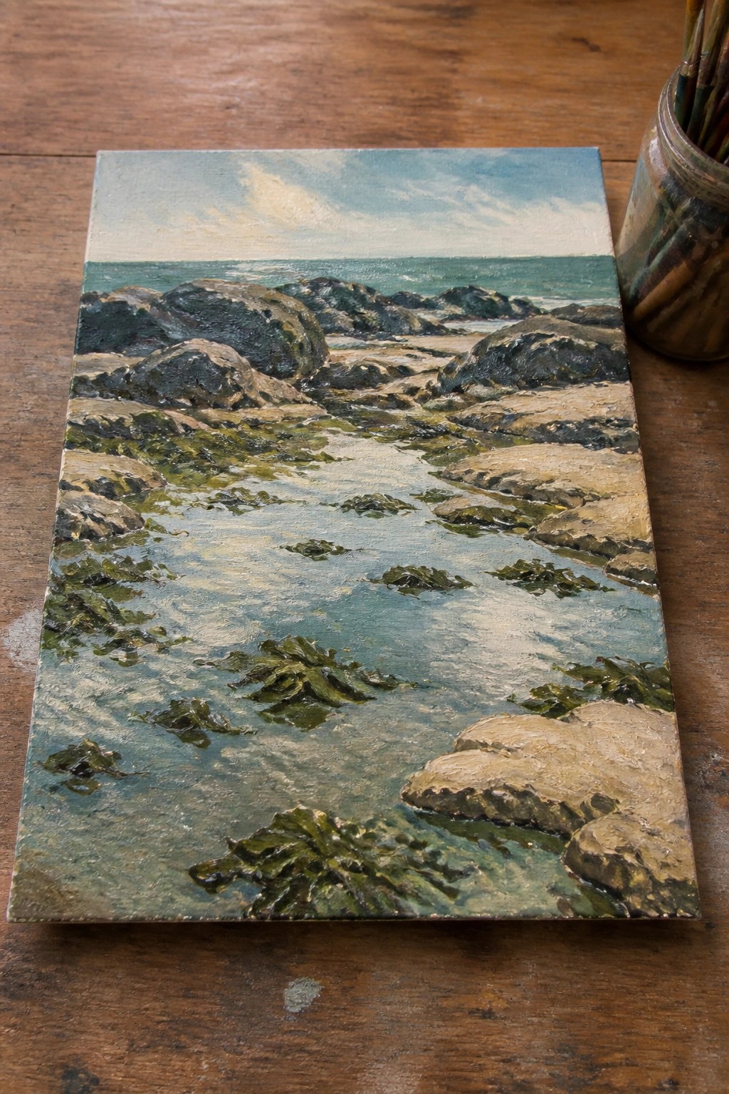 Seascape painting of rocky shore with seaweed in clear water and distant waves beside brushes.