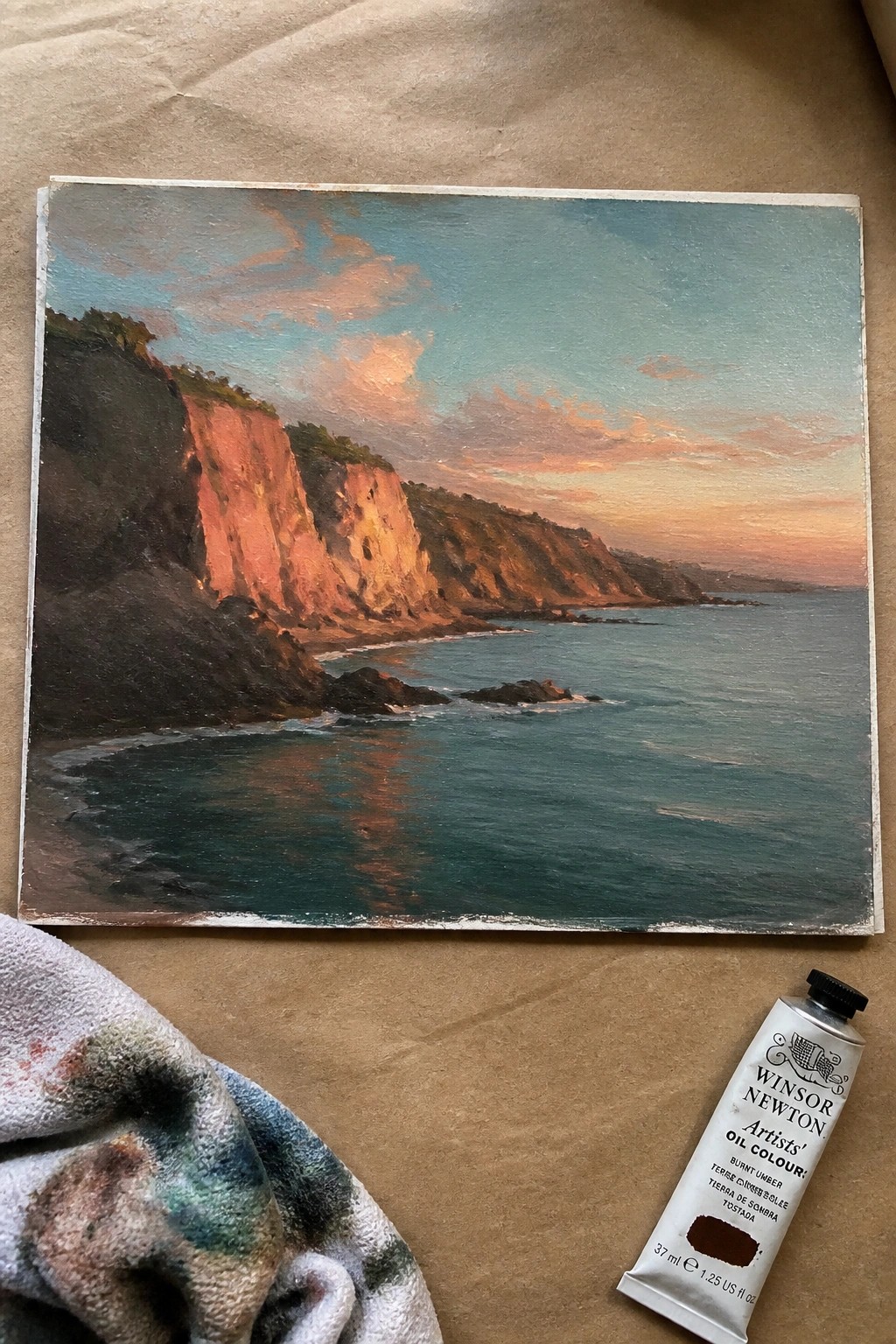 Oil painting of sunset cliffs by sea beside burnt umber paint tube and rag