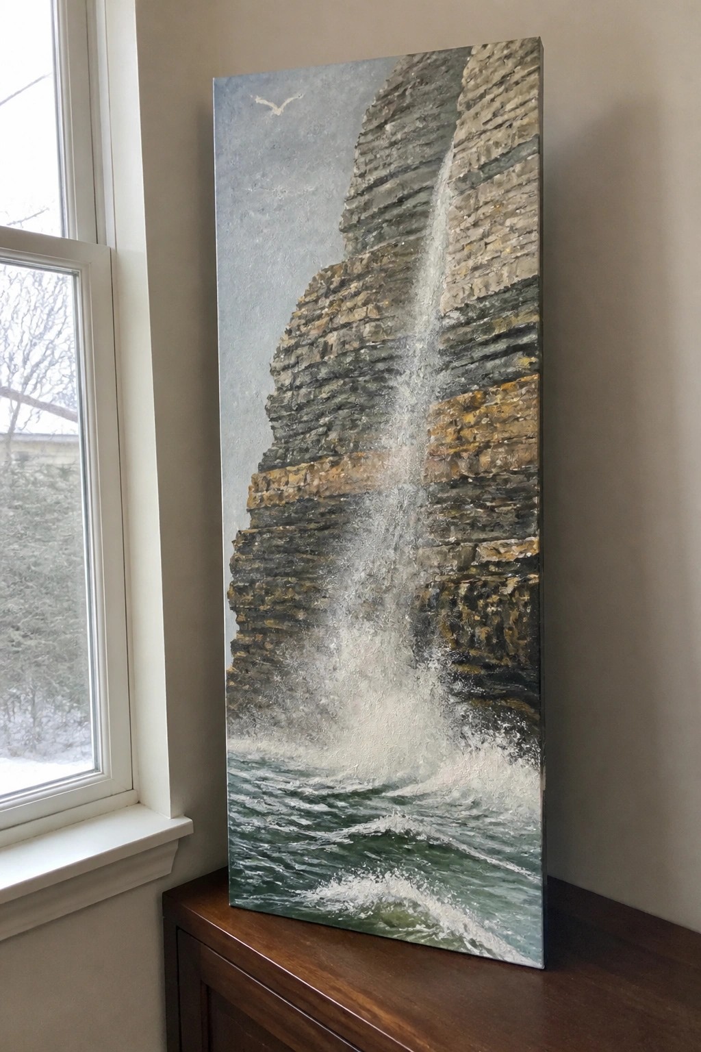 Realistic painting of waterfall cascading down rocky cliffs into green ocean waves beside window