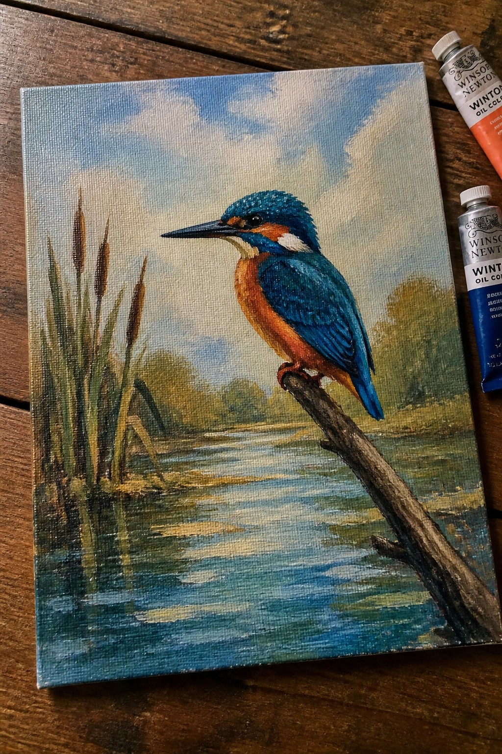 Colorful oil painting of kingfisher on branch over water beside paint tubes.