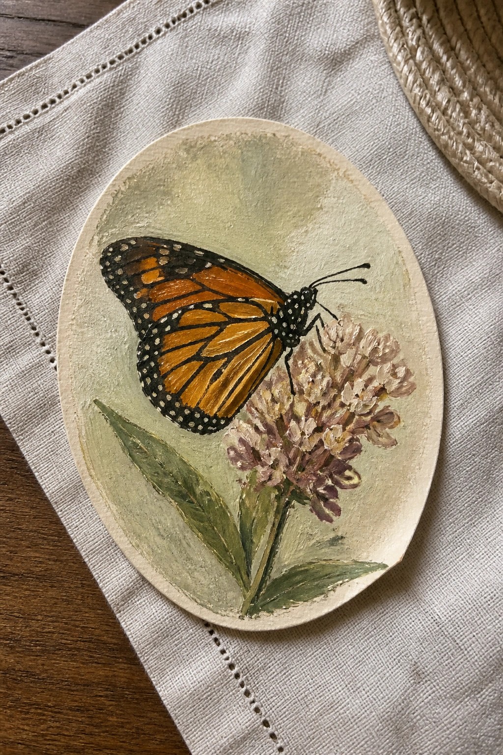 Oval wooden plaque with orange monarch butterfly on purple flowers atop beige fabric.