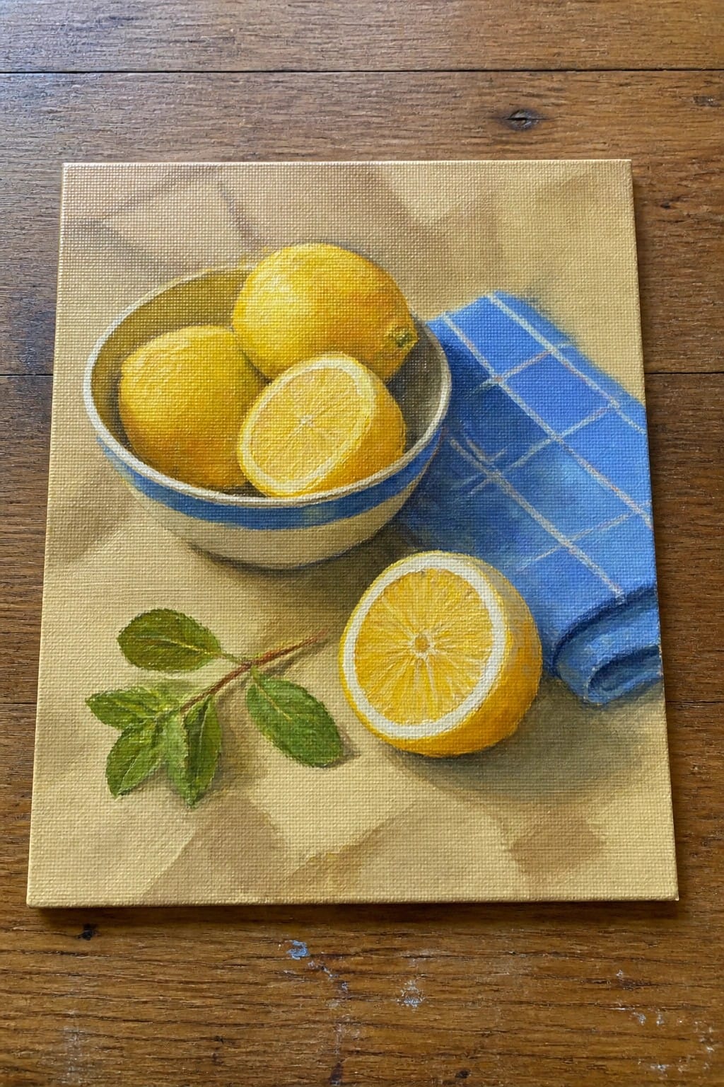 Yellow lemons in striped bowl beside blue checkered cloth, sliced lemon, and leaves