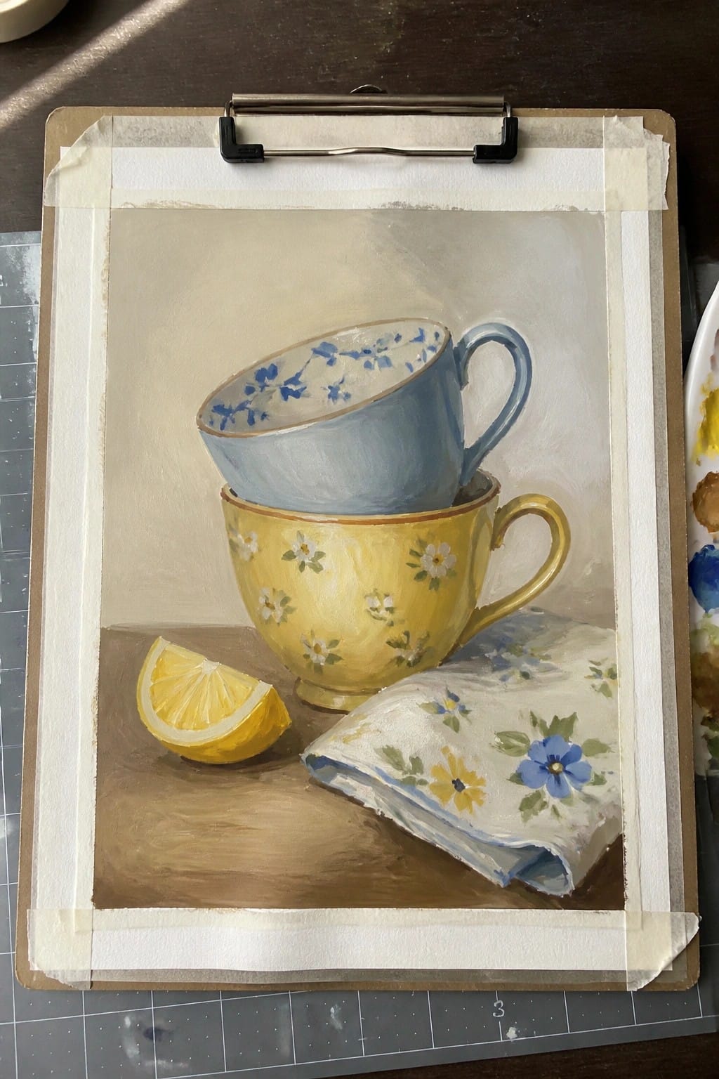 Still life painting of stacked yellow and blue floral teacups beside lemon and napkin