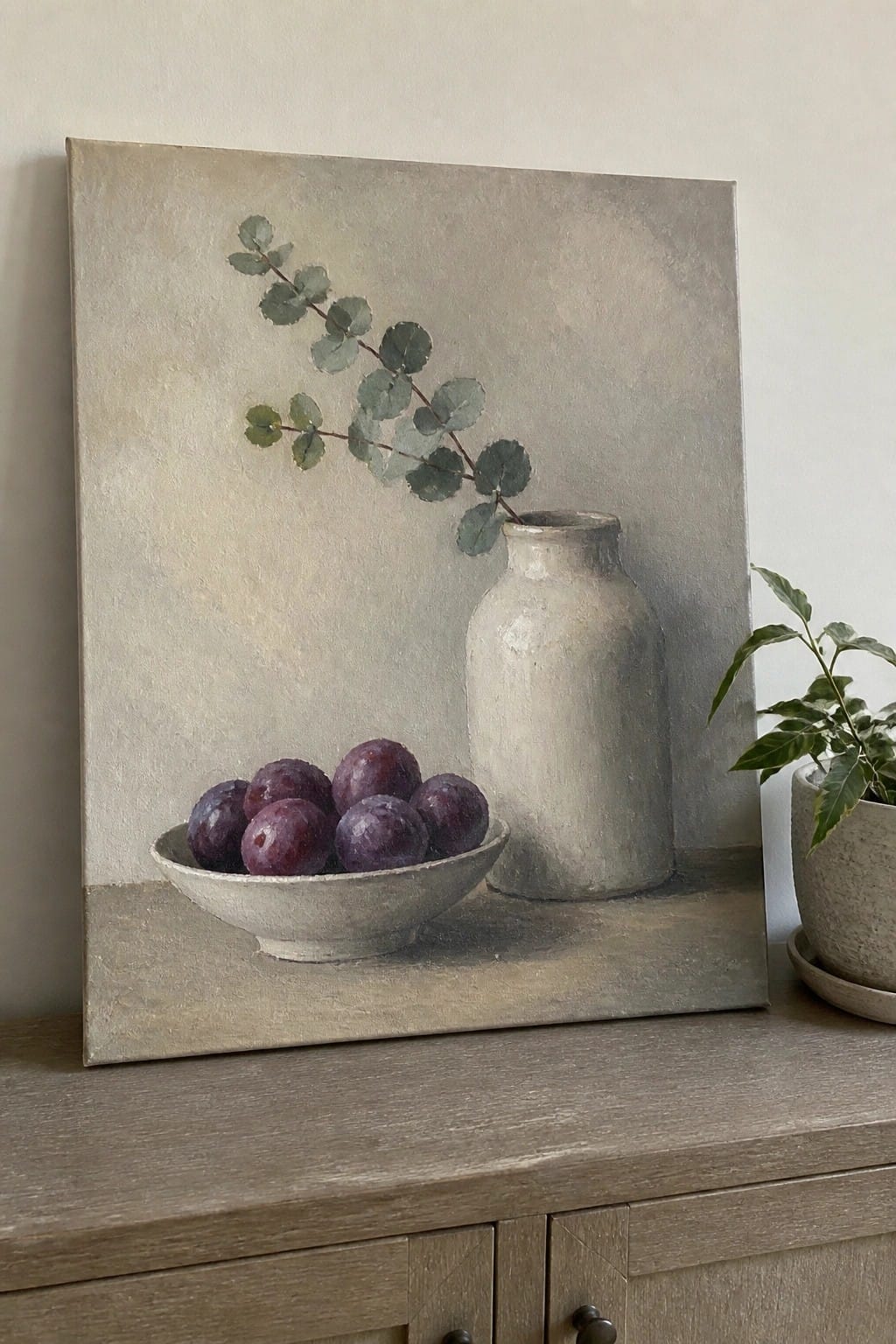 Still life painting of purple plums in bowl beside white vase with eucalyptus branch