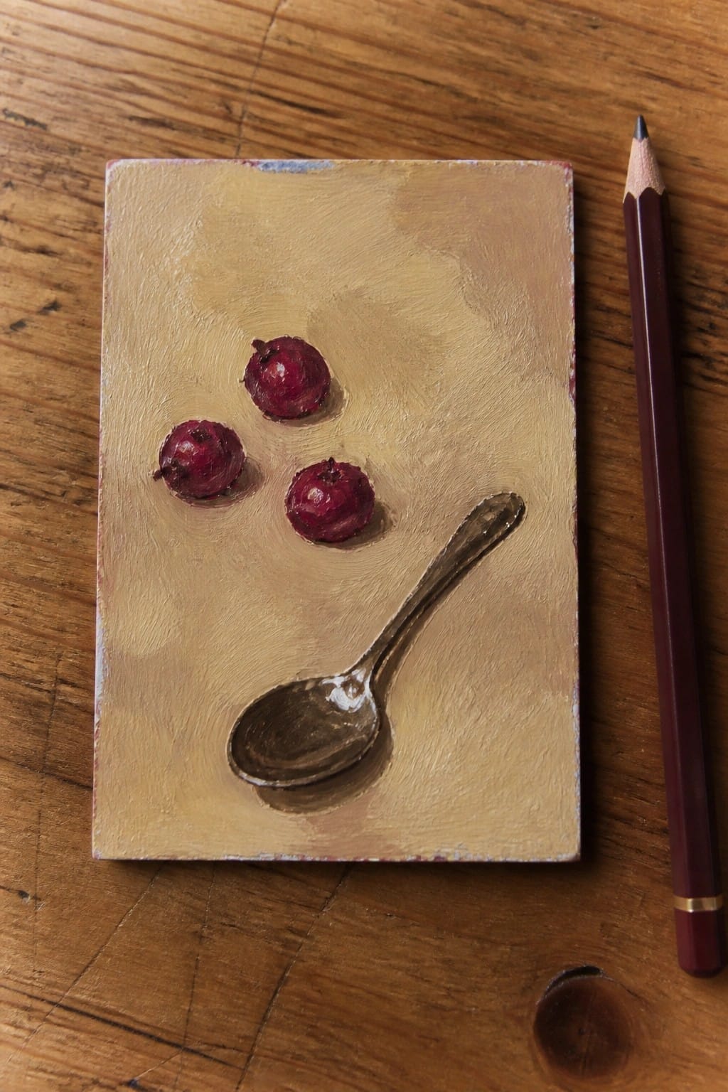 An oil painting of three small red berries and a spoon on a textured beige background next to a pencil on wood.