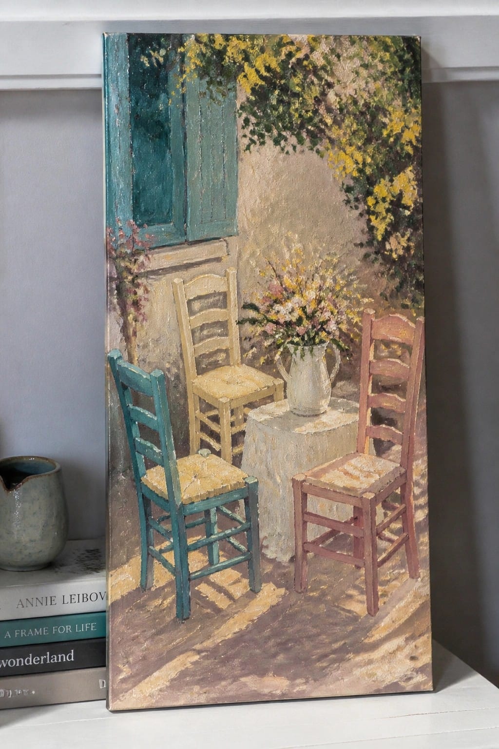 Impressionist painting of three colorful chairs around a table with floral pitcher by blue shutters