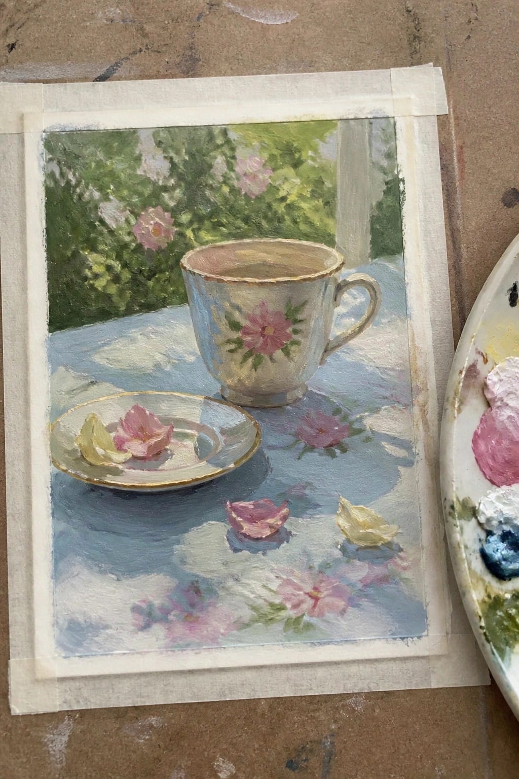 An oil painting of a floral teacup and saucer on a blue table with scattered pink and yellow petals, set against a green garden background with pink roses.