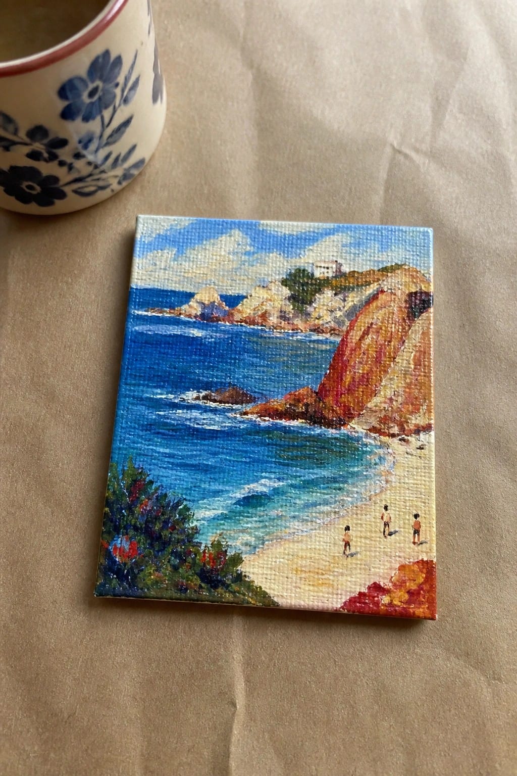 Vibrant miniature painting of beach with blue sea, colorful cliffs, tiny figures beside floral mug