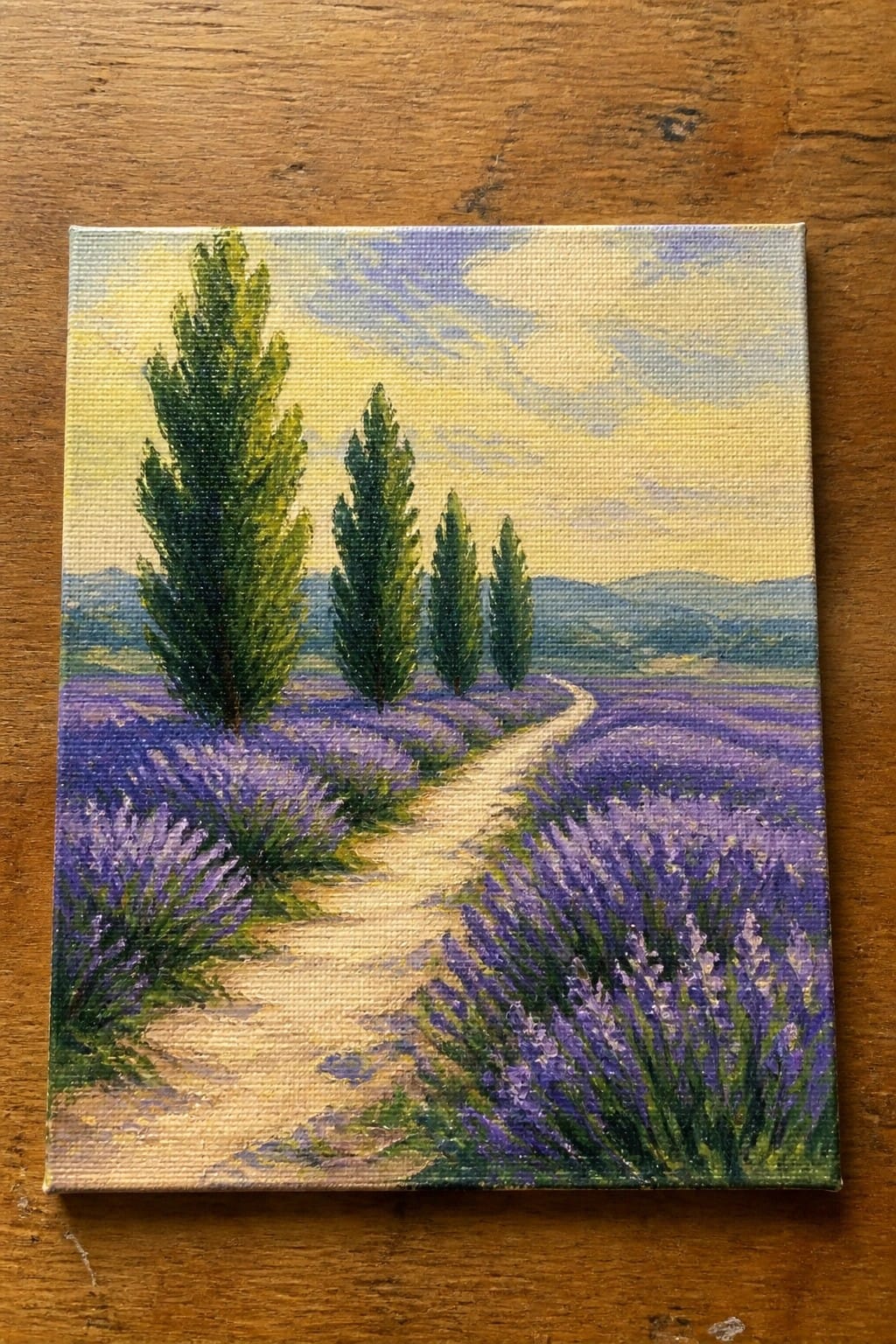Small canvas painting of lavender field with winding path, cypress trees, and hills on wood.