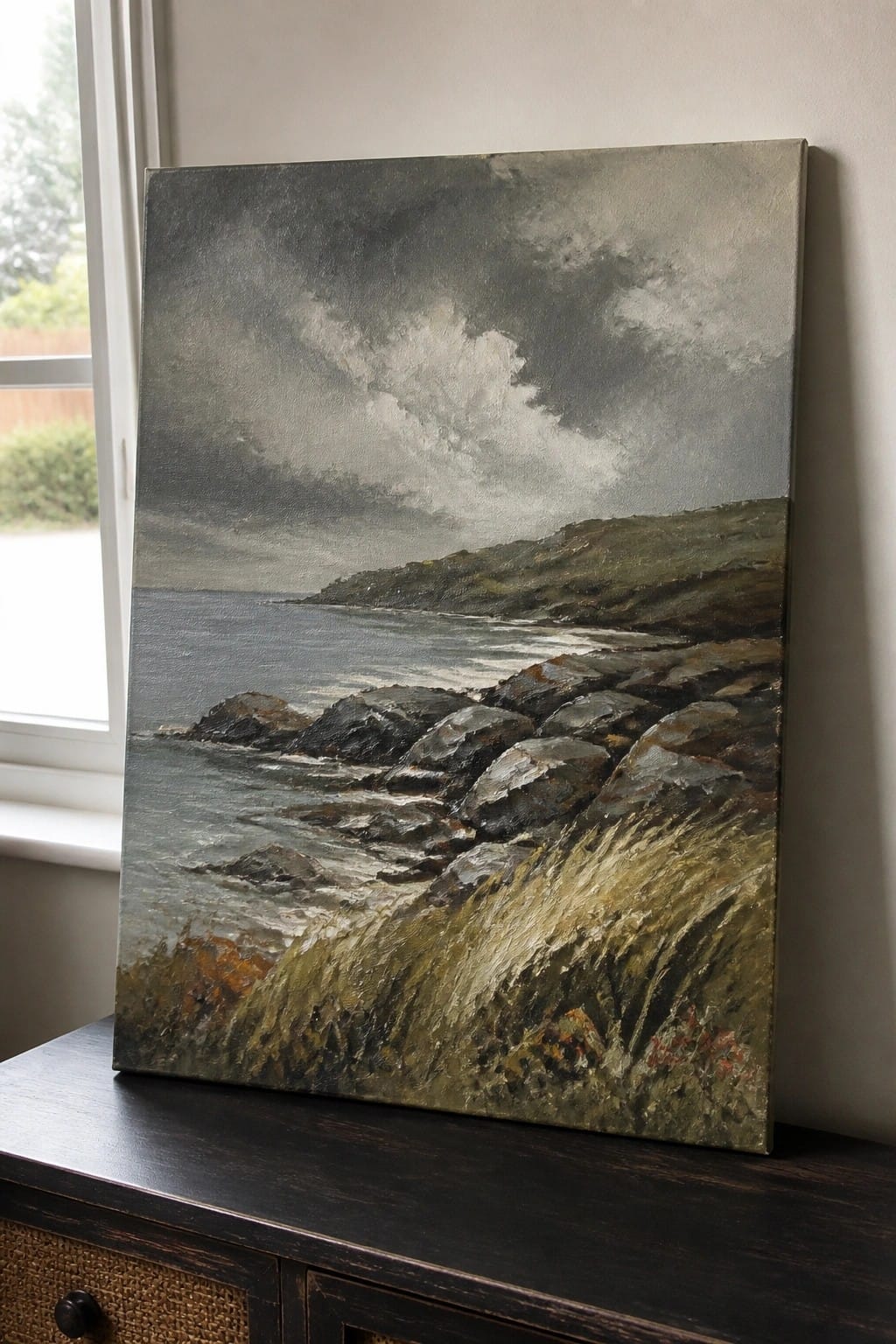 Stormy seascape painting with rocks and waves on dark wooden cabinet beside window