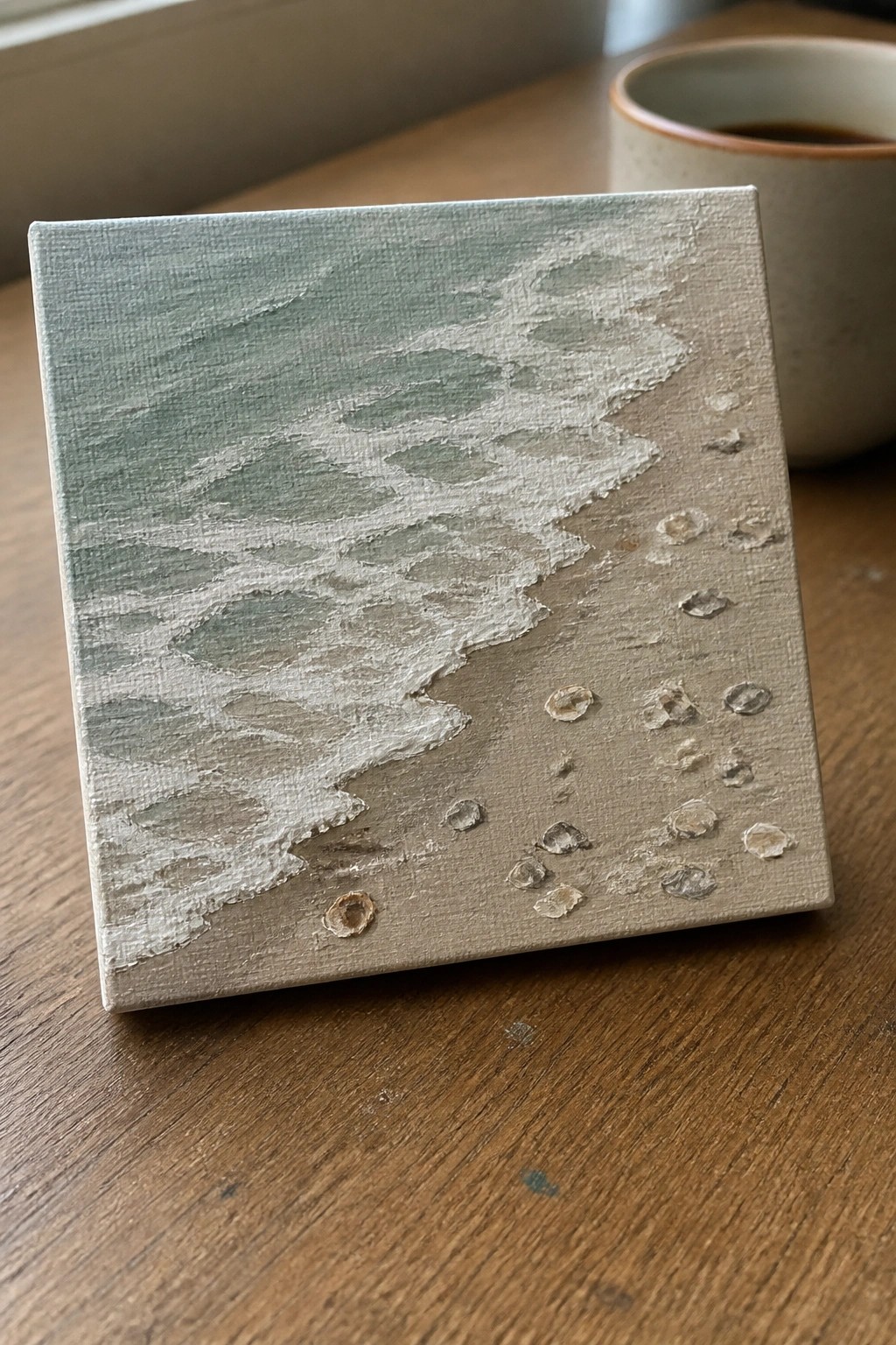 Canvas painting of green waves on textured beige sand beside ceramic cup on wood table