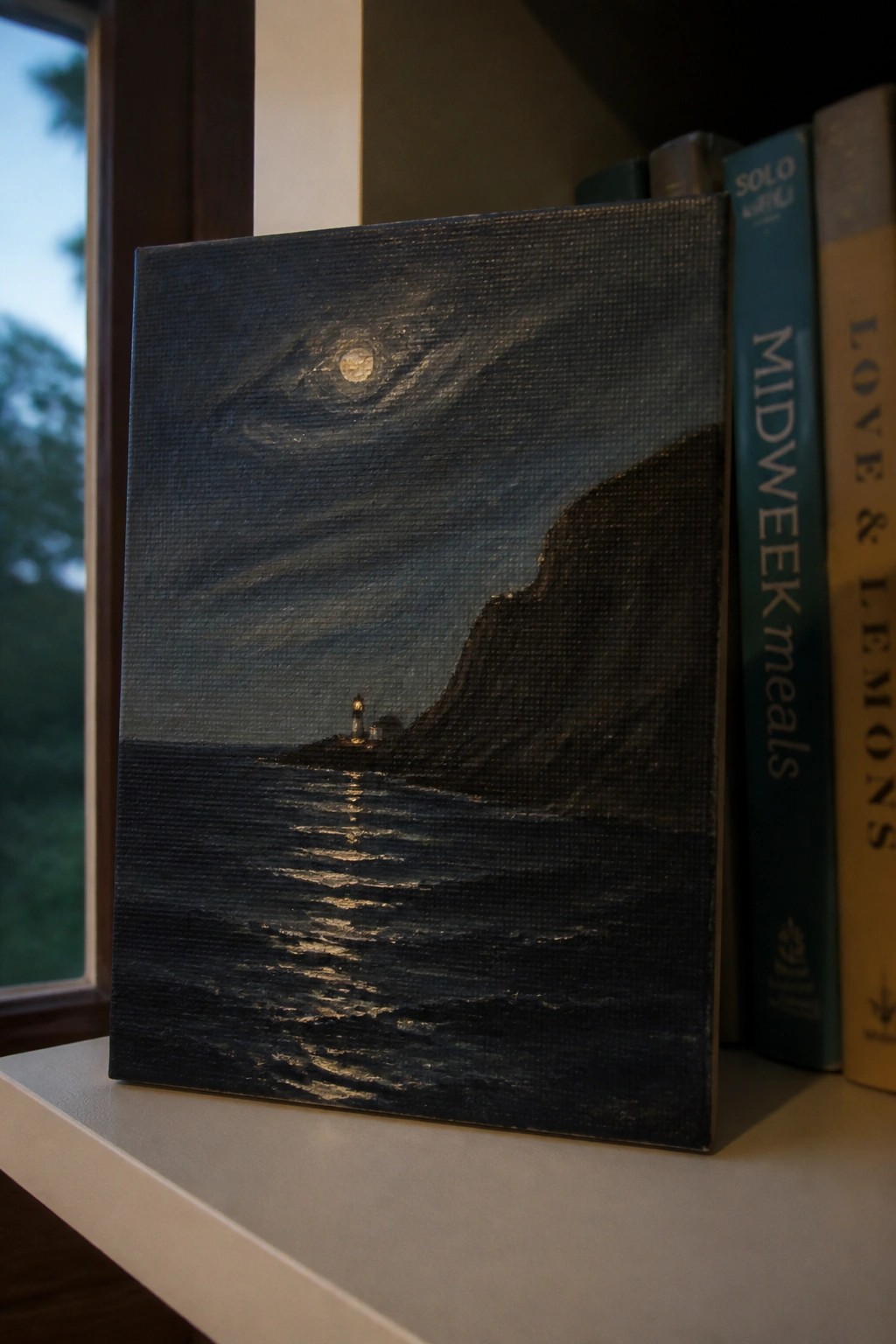 Painted canvas of moonlit sea with lighthouse leaning against books on shelf