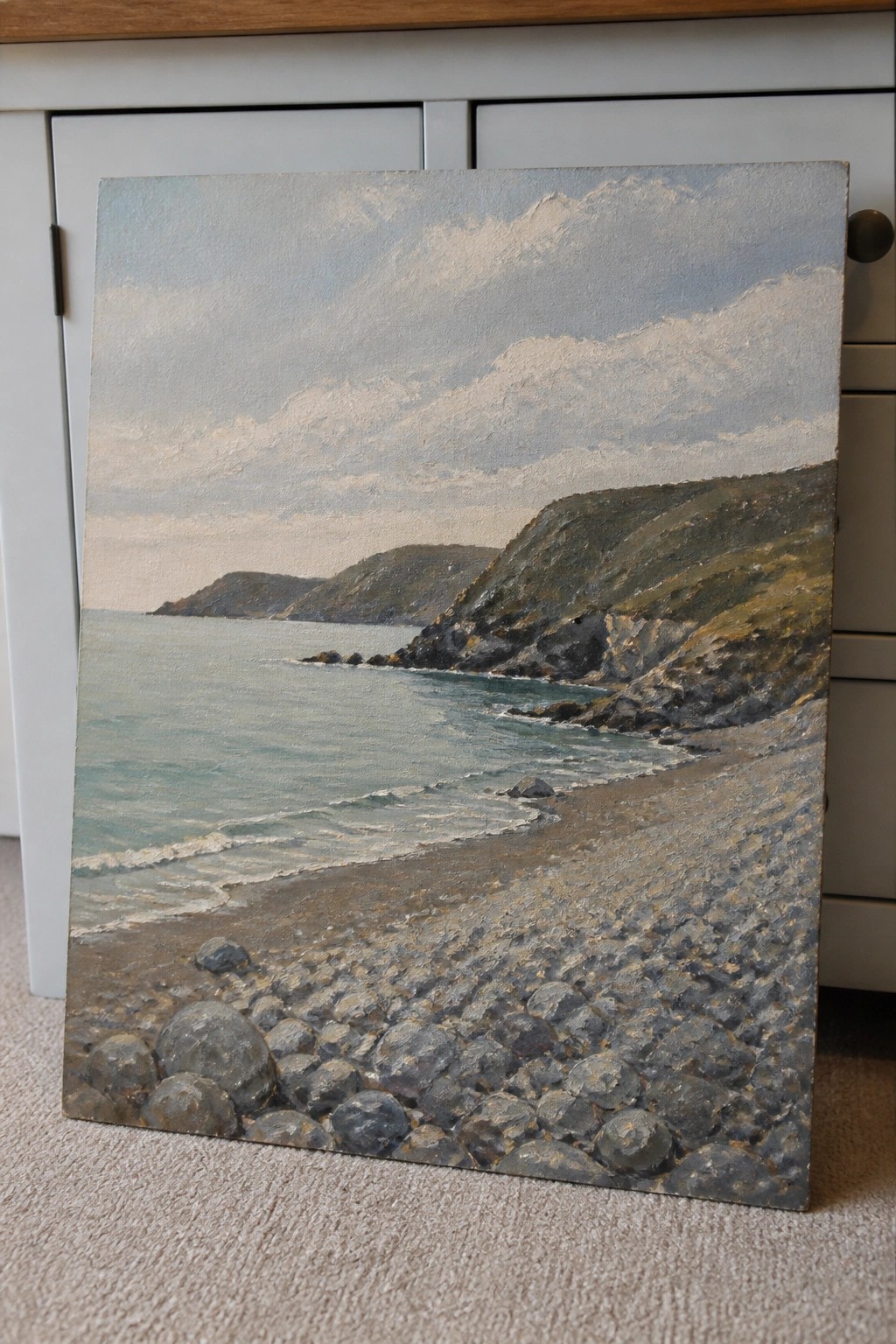 Landscape painting of pebble beach, calm sea, rocky cliffs and cloudy mountains.