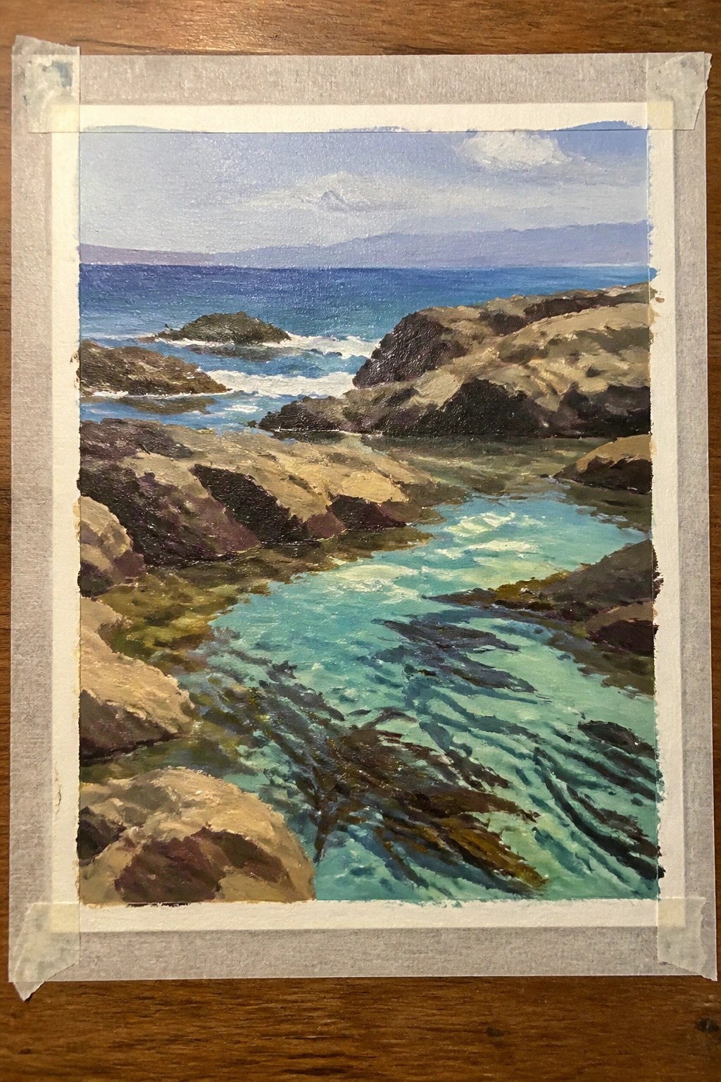 Rocky coastline painting with turquoise waves, dark rocks, and distant mountains under blue sky