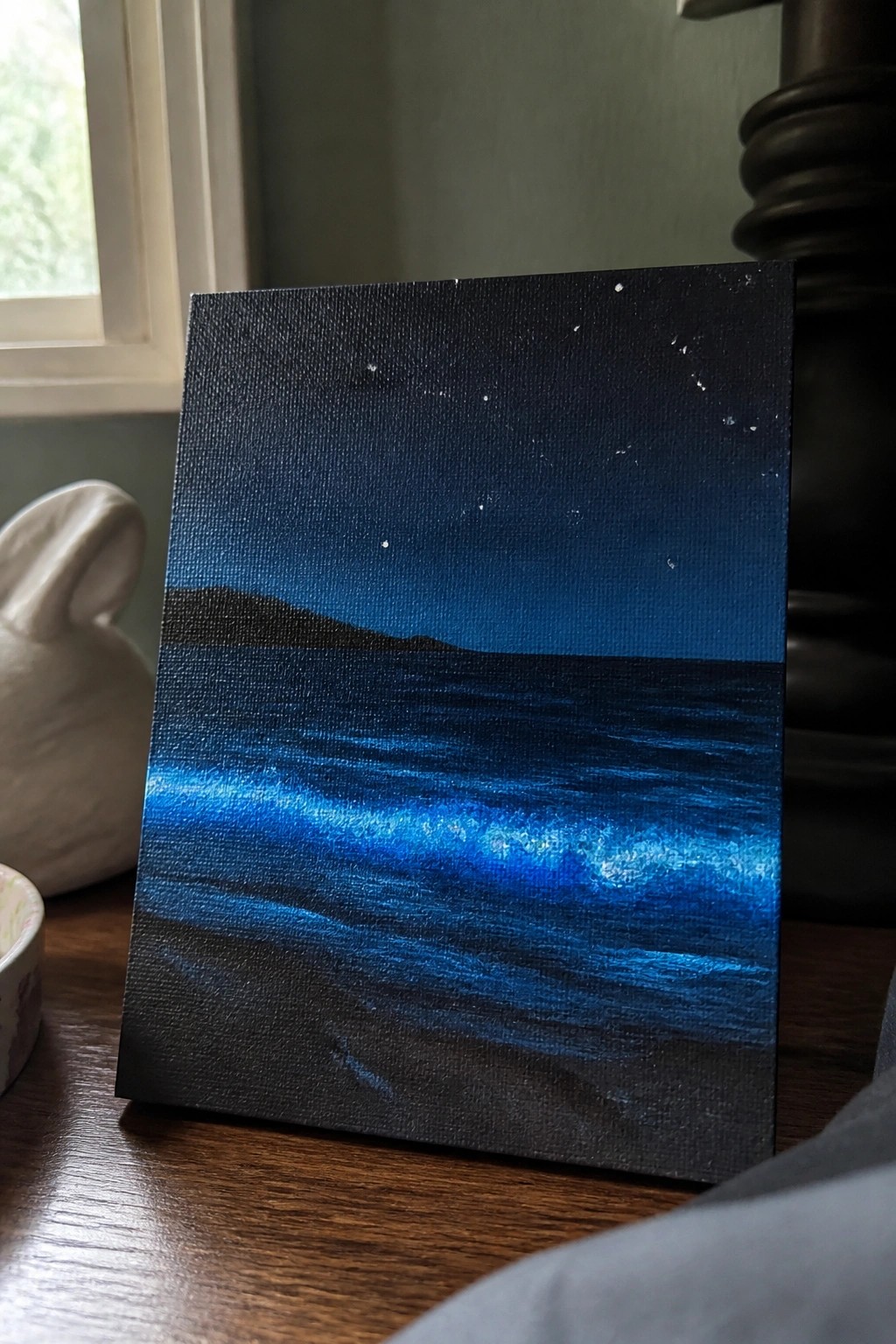 A small oil painting on canvas showing a starry night sky over dark water with glowing blue waves and a black foreground.