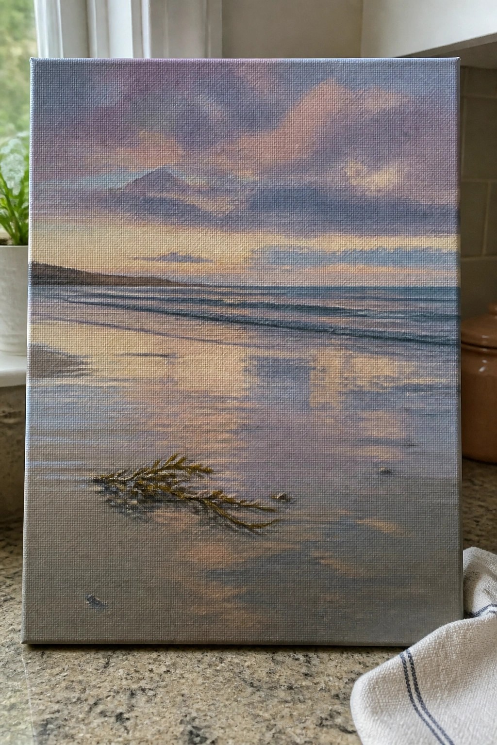 Seascape painting with purple-pink sky, mountains, calm water, and seaweed on sand, on countertop.