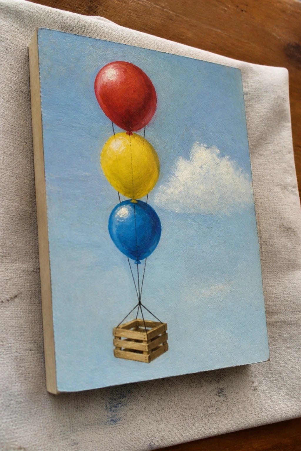 Red, yellow, and blue balloons lifting a wooden crate in a blue sky painting on canvas